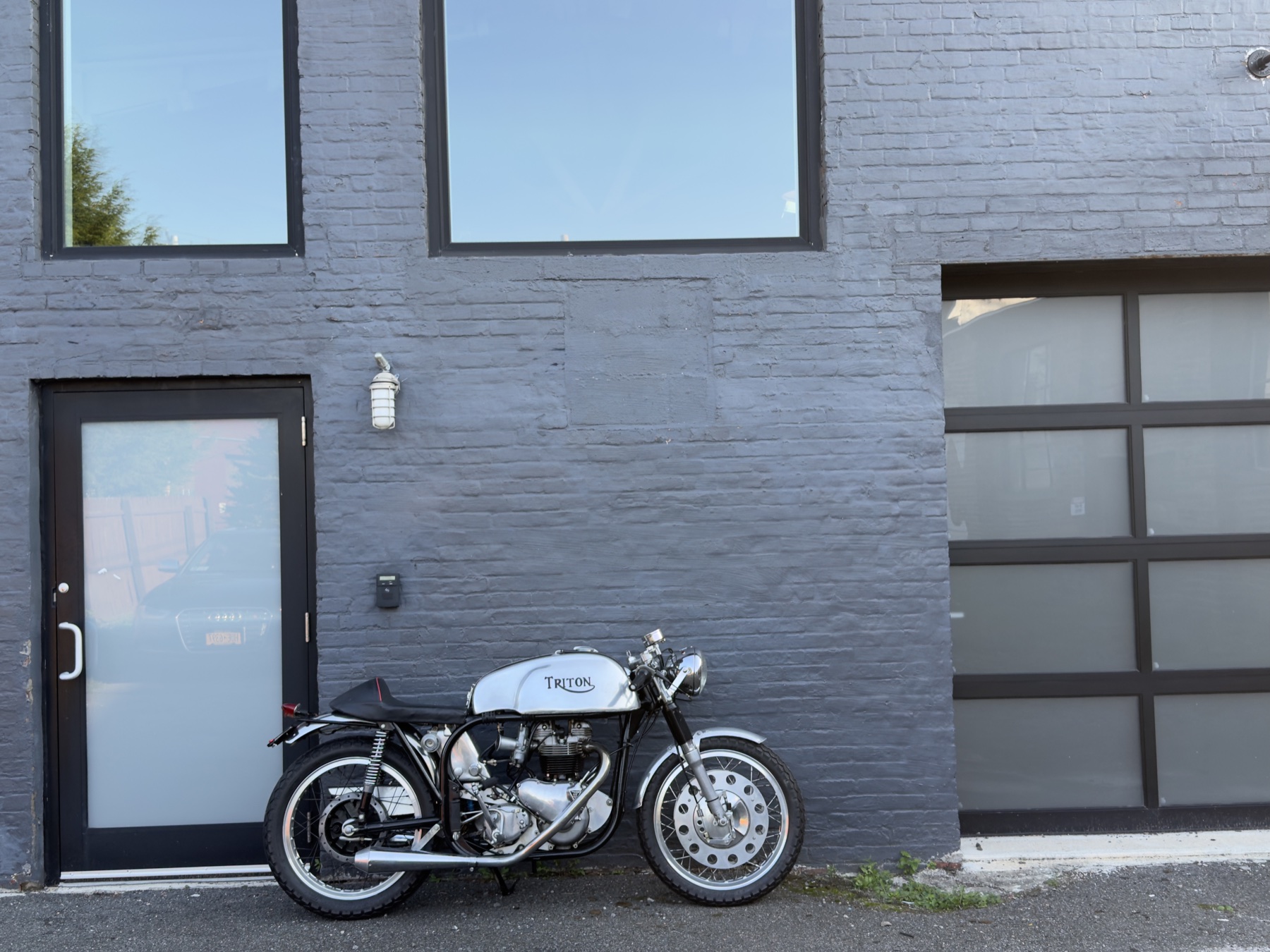A Triton café racer parked against the gray brick beside the drive-in bay